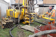 Ein Arbeiter in orangefarbener Arbeitskleidung bedient eine große Bohrmaschine auf einem Grundstück. Neben ihr liegen Rohre, und es gibt eine rote Abfallmulde sowie Schläuche im Umfeld. Im Hintergrund sind Wände und ein Zaun sichtbar.