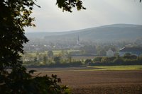 Eine ländliche Landschaft mit sanften Hügeln im Hintergrund. Im Vordergrund gibt es ein Feld, während im Hintergrund eine Ortschaft mit Wohnhäusern und einer Kirche sichtbar ist. Der Himmel ist leicht bewölkt und die Umgebung wirkt ruhig und friedlich.