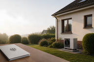 Eine moderne Wärmepumpe steht an der Wand eines Hauses in einem grünen Garten. Im Vordergrund liegt ein offenes Heft auf einem Tisch. Der Himmel ist hell und die Umgebung wirkt ruhig und einladend.
