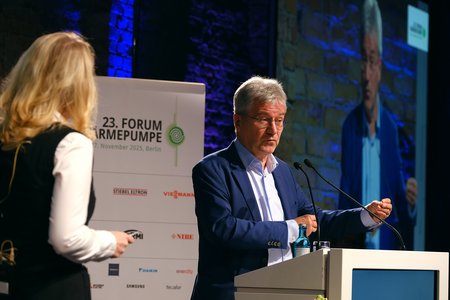 Ein Mann steht an einem Rednerpult und spricht bei einem Forum. Eine Frau steht neben ihm und hört zu. Im Hintergrund ist ein Bildschirm mit einer Präsentation zu sehen. Die Veranstaltung findet am 23. November 2015 in Berlin statt.