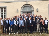 Eine Gruppe von etwa 40 Personen steht treppabwärts vor einem Gebäude. Die Teilnehmer sind in formeller und legerer Kleidung gekleidet und lächeln in die Kamera. Hinter ihnen ist eine fassadenartige Wand mit einem großen Fenster und zwei seitlichen Lampen zu sehen.