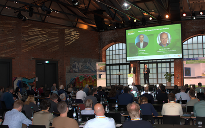 Eine Konferenzveranstaltung in einem Industriegebäude. Auf der Bühne stehen zwei Redner, während das Publikum im Hintergrund zuhört. An einer Wand sind Grafiken und Präsentationsmaterial sichtbar. Die Atmosphäre ist professionell und aufmerksam.