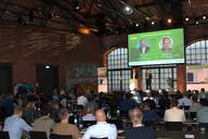 Eine Konferenzveranstaltung in einem Industriegebäude. Auf der Bühne stehen zwei Redner, während das Publikum im Hintergrund zuhört. An einer Wand sind Grafiken und Präsentationsmaterial sichtbar. Die Atmosphäre ist professionell und aufmerksam.