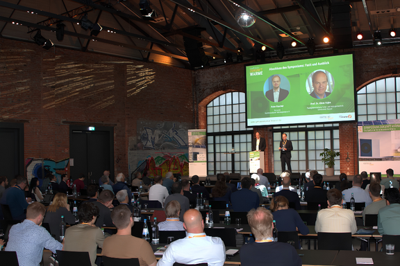 Eine Konferenzveranstaltung in einem Industriegebäude. Auf der Bühne stehen zwei Redner, während das Publikum im Hintergrund zuhört. An einer Wand sind Grafiken und Präsentationsmaterial sichtbar. Die Atmosphäre ist professionell und aufmerksam.
