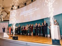 Eine Gruppe von Menschen steht auf einer Bühne und applaudiert. Im Hintergrund wird eine Präsentation mit der Aufschrift „Wohnungen sollen seriell auf Net-Zero-Standard saniert werden“ gezeigt. Funkenregen entsteht, was eine feierliche Atmosphäre schafft.