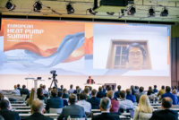 Eine Konferenz zum Thema Wärmepumpen findet in Nürnberg statt. Auf der Bühne spricht ein Referent, während auf einem großen Bildschirm ein weiterer Experte aus der Ferne zugeschaltet ist. Im Publikum sitzen viele Teilnehmer, die auf das Geschehen achten.