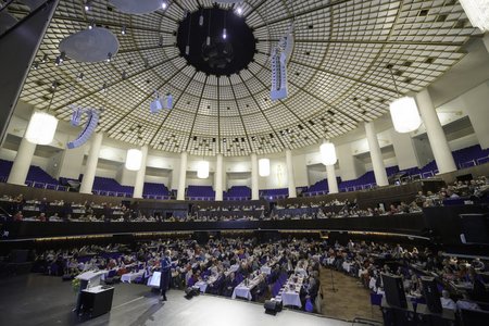 Ein großer Konferenzraum mit einer hohen, gewölbten Decke und vielen Sitzplätzen. Zahlreiche Tische sind besetzt, während Zuhörer einer Präsentation auf der Bühne folgen. Die Atmosphäre wirkt geschäftig und konzentriert.