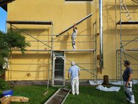 Drei Arbeiter stehen auf einem Gerüst vor einer gelben Hauswand. Einer trägt ein langes Bauteil und steht auf einer höheren Ebene, während die anderen ihn beobachten. Der Boden ist mit Materialien für die Arbeit bedeckt, und im Hintergrund ist ein Fenster sichtbar.