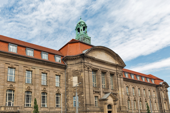Historisches Gebäude mit herausragender Architektur, einschließlich einem großen Eingang und einem Glockenturm. Die Fassade ist mit symmetrischen Fenstern und einem roten Ziegeldach gestaltet. Der Himmel zeigt einige Wolken.