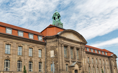 Historisches Gebäude mit herausragender Architektur, einschließlich einem großen Eingang und einem Glockenturm. Die Fassade ist mit symmetrischen Fenstern und einem roten Ziegeldach gestaltet. Der Himmel zeigt einige Wolken.