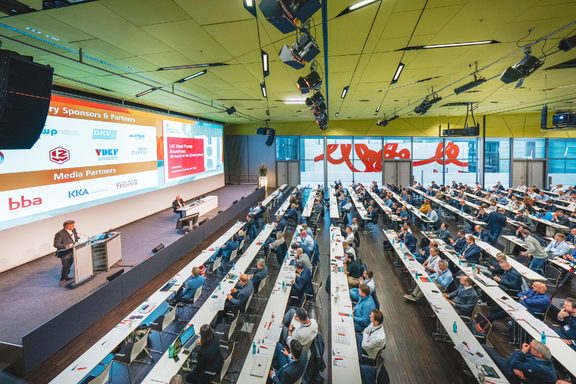 Eine Konferenz mit einem Redner auf der Bühne, der vor einem Publikum spricht. Die Teilnehmer sitzen in Reihen an Tischen, während Präsentationen auf einer großen Leinwand gezeigt werden. Der Raum ist gut beleuchtet und modern eingerichtet.