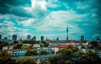 Das Bild zeigt eine Stadtansicht mit modernen Hochhäusern und einer Vielzahl von Bäumen. Im Hintergrund ragt der Fernsehturm von Berlin in den Himmel, umgeben von einer dramatischen Wolkenlandschaft. Die Farben wirken lebhaft und einladend.
