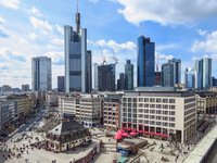 Das Bild zeigt eine Stadtansicht von Frankfurt am Main mit modernen Wolkenkratzern und der belebten Innenstadt. Im Vordergrund sieht man ein historisches Gebäude und Plätze mit zahlreichen Menschen und Sitzgelegenheiten. Der Himmel ist blau mit einigen Wolken.