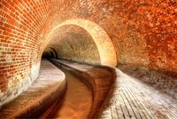 Eine unterirdische Ziegelsteinstruktur mit gewölbter Decke, die einen schmalen, gewundenen Gang zeigt. Die Wände bestehen aus rotbraunen Ziegeln, die eine warme Atmosphäre schaffen. Am Boden verläuft ein schmaler Kanal, der in die Tiefe führt.