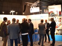 Eine Gruppe von Fachleuten steht um einen Stehtisch auf einer Messe. Sie diskutieren zum Thema erneuerbare Energien. Im Hintergrund sind Informationsstände mit Grafiken und Texten über nachhaltige Energien zu sehen.