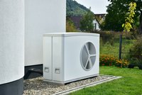 Eine weiße Wärmepumpe steht an der Wand eines Hauses, umgeben von Kies und Gras. Im Hintergrund sind undeutlich Gartenpflanzen und ein Zaun sichtbar. Die Wärmepumpe hat eine runde Belüftung auf der Vorderseite und ist eine moderne Heiz- und Kühltechnologie.