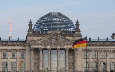 Das Bild zeigt die Fassade des Reichstagsgebäudes in Berlin mit einer markanten Glaskuppel. Vor dem Gebäude weht die deutsche Flagge. Es ist ein klarer Himmel im Hintergrund zu sehen.