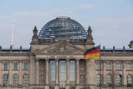Das Bild zeigt die Fassade des Reichstagsgebäudes in Berlin mit einer markanten Glaskuppel. Vor dem Gebäude weht die deutsche Flagge. Es ist ein klarer Himmel im Hintergrund zu sehen.