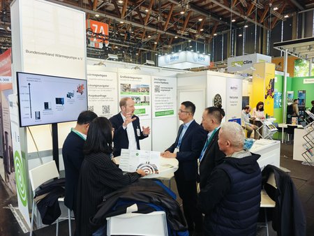 Auf der Messe sind mehrere Personen an einem Stand versammelt. Ein Mann präsentiert Informationen auf einem Bildschirm, während andere Zuhörer ihm aufmerksam zuhören. Der Stand ist mit Grafiken und Informationen zum Thema Wärmepumpen dekoriert.