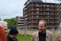 Eine Gruppe von Männern steht im Vordergrund, während im Hintergrund ein Gebäude im Bau zu sehen ist, das mit Gerüsten versehen ist. Die Umgebung ist grün, mit Bäumen und hohem Gras. Der Himmel ist bewölkt.