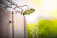 Eine Duschkopf aus Edelstahl strahlt Wasser in einem hellen, lichtdurchfluteten Badezimmer. Im Hintergrund ist unscharfe, grüne Vegetation zu sehen. Der Fokus liegt auf dem Wasser, das gleichmäßig herabfließt.