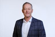 Ein mittelalter Mann mit kurzen, grauen Haaren und einem freundlichen Gesichtsausdruck trägt einen blauen Blazer über einem weißen Hemd. Er steht vor einem weißen Hintergrund und schaut direkt in die Kamera.