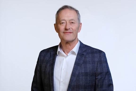 Ein mittelalter Mann mit kurzen, grauen Haaren und einem freundlichen Gesichtsausdruck trägt einen blauen Blazer über einem weißen Hemd. Er steht vor einem weißen Hintergrund und schaut direkt in die Kamera.