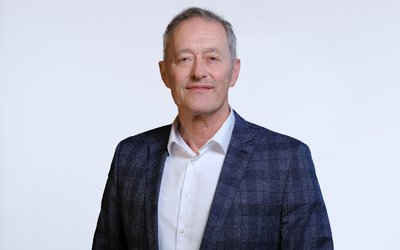 Ein mittelalter Mann mit kurzen, grauen Haaren und einem freundlichen Gesichtsausdruck trägt einen blauen Blazer über einem weißen Hemd. Er steht vor einem weißen Hintergrund und schaut direkt in die Kamera.