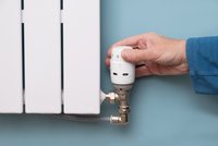 Eine Hand dreht einen Thermostatkopf an einem Heizkörper. Der Heizkörper ist weiß und an einer blauen Wand montiert. Der Thermostatkopf ist rund und hat eine digitale Anzeige.