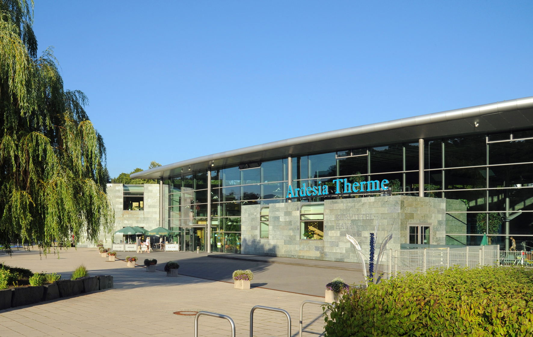 Die Ardesia Therme ist ein modernes Gebäude mit großen Glasfenstern und einer einladenden Fassade. Im Vordergrund sind Pflanzenkübel zu sehen. Die entspannende Atmosphäre der Therme ist durch das sonnige Wetter und die klare blaue Himmel deutlich spürbar.